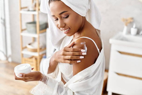 Femme afro-américaine en peignoir avec des tresses afro tenant pot de crème dans la salle de bain moderne. - Photo, image
