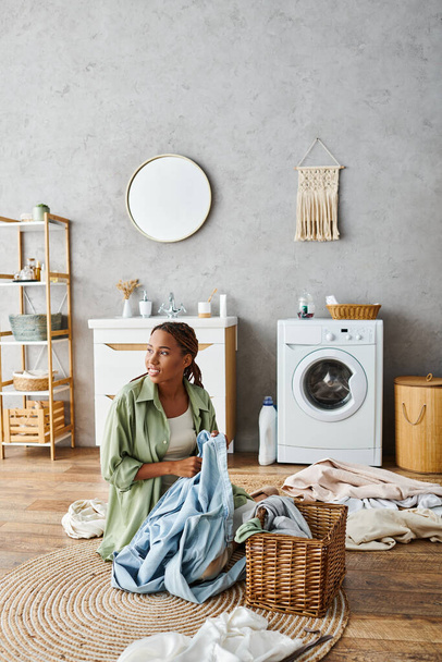 Uma mulher afro-americana com tranças afro sentada no chão em frente a uma máquina de lavar roupa, lavando a roupa em um banheiro. - Foto, Imagem