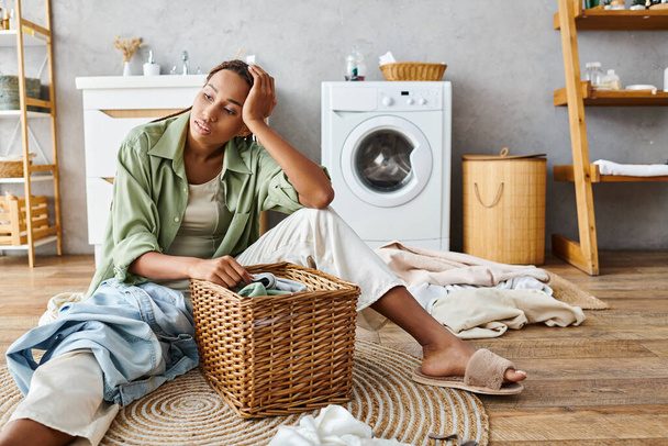 Африканська американка з афро косами сидить поруч з кошиком для білизни у ванній кімнаті, готуючись до прання одягу. - Фото, зображення