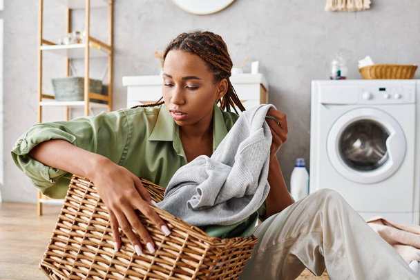 Afroamerykanka z afro warkoczami organizuje pranie w łazience, siedząc na podłodze z koszem na pranie. - Zdjęcie, obraz