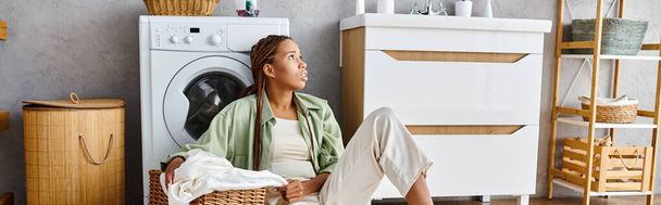 Mulher afro-americana com tranças afro senta-se na frente de uma máquina de lavar roupa, lavando roupa em um banheiro. - Foto, Imagem