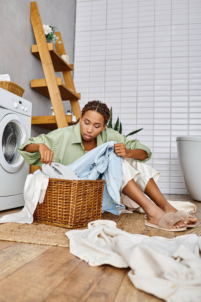 Una mujer afroamericana con trenzas afro sentada cerca de una lavadora, comprometida en la tarea de lavar la ropa en un baño. - Foto, Imagen