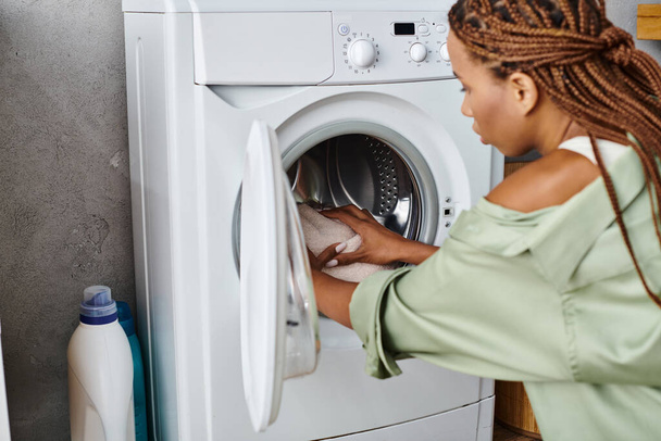 African American woman with afro braids putting clothes into a dryer in a bathroom while doing laundry. - Photo, Image