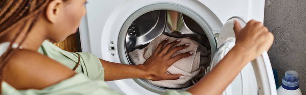 Uma mulher afro-americana com tranças limpa diligentemente a frente de uma máquina de lavar roupa em um banheiro. - Foto, Imagem