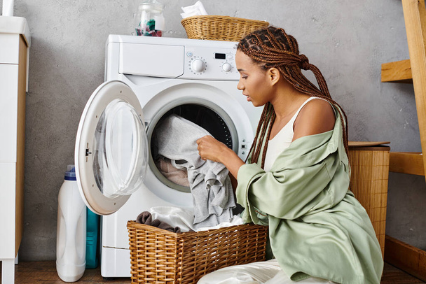 African American woman with afro braids sitting by a washing machine doing laundry in a bathroom. - Fotografie, Obrázek