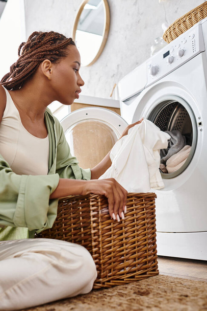 Eine afroamerikanische Frau mit Afro-Zöpfen sitzt vor einer Waschmaschine und wäscht Wäsche im Badezimmer. - Foto, Bild