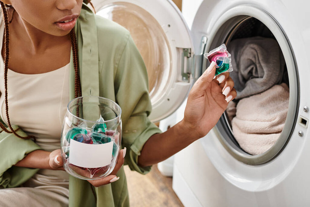 Afroameričanka s afro copánky si užívá klidného okamžiku, drží kapsle s gelem vedle pračky v útulné koupelně. - Fotografie, Obrázek