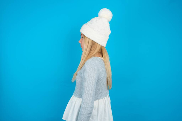Hermosa niña con sombrero de punto blanco y suéter azul mirando a un lado, pose de perfil relajante con cara natural con sonrisa confiada. - Foto, Imagen