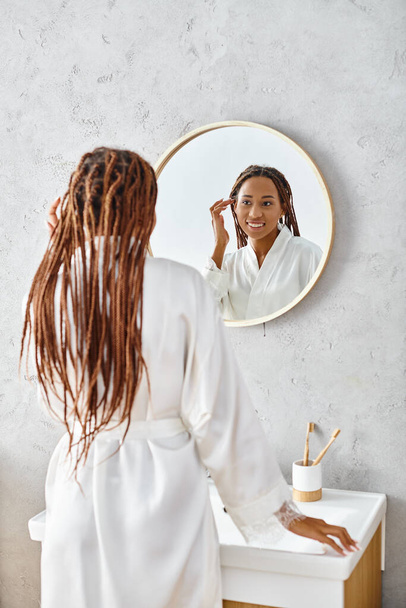 Una mujer afroamericana con trenzas afro se cepilla el pelo en un baño moderno, centrándose en la belleza y la higiene. - Foto, imagen