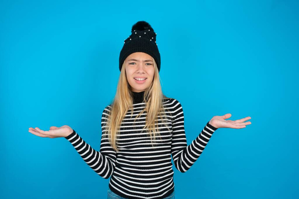 Indignant beautiful kid girl wearing knitted black hat and striped turtleneck over blue background gestures in bewilderment, frowns face with dissatisfaction. - Photo, Image