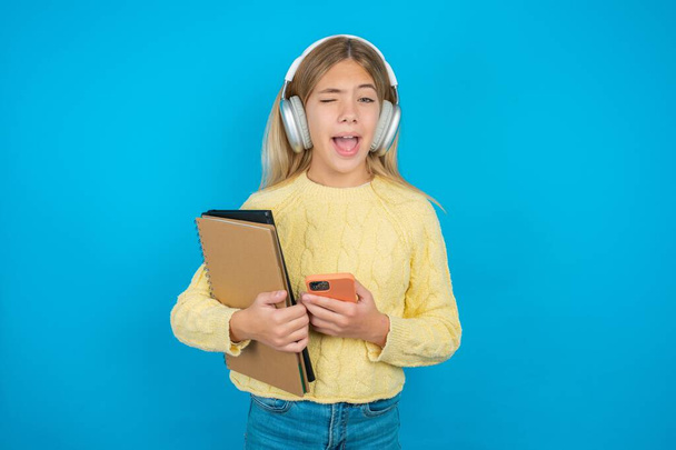 Excited Beautiful kid girl wearing yellow sweater winking and eye hold smart phone use read social network news - Photo, Image