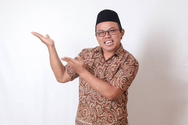 Portré izgatott ázsiai férfi batik ing és songkok mosolygott, és néz a kamerába mutató két kézzel és ujjal az oldalán. Izolált kép szürke háttérrel - Fotó, kép