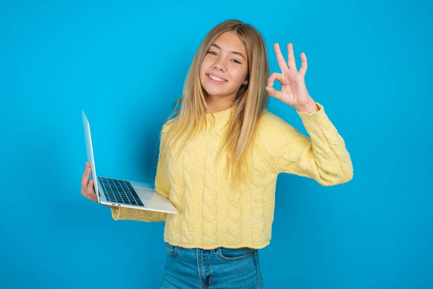 Pozitív Gyönyörű gyerek lány visel sárga pulóver tartsa vezeték nélküli netbook kéz ujjak mutatják okey szimbólum - Fotó, kép