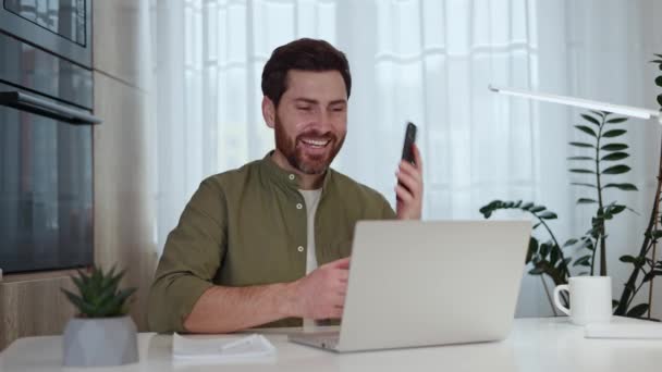 Vidám fehér férfi veszi fel a bejövő telefont, miközben laptopon dolgozik az otthoni munkahelyén. Jóképű fickó folytatja a munkát, miközben személyes beszélgetést folytat. A többfunkciós feladatok fogalma. - Felvétel, videó