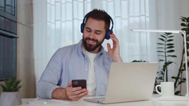 Elégedett távoli tervező fülhallgató zenél a mobil eszközön, miközben munkanap kezdődik a számítógépen. A fejlett munkavállaló a termelékenység növelése és a folyamatokra való összpontosítás érdekében pályákat használ. - Felvétel, videó