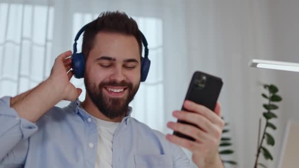 Caucasian young man listening to music in wireless headphones connected to phone and moving relaxed indoor. Attractive male choosing soundtrack from playlist on modern gadget and nodding head to beat. - Footage, Video