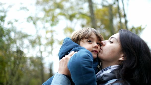 Moeder tillen op in armen en kussen in wang staan buiten tijdens de herfst herfst seizoen, authentieke levensstijl familie scène van moeder en kind liefdevolle relatie - Foto, afbeelding