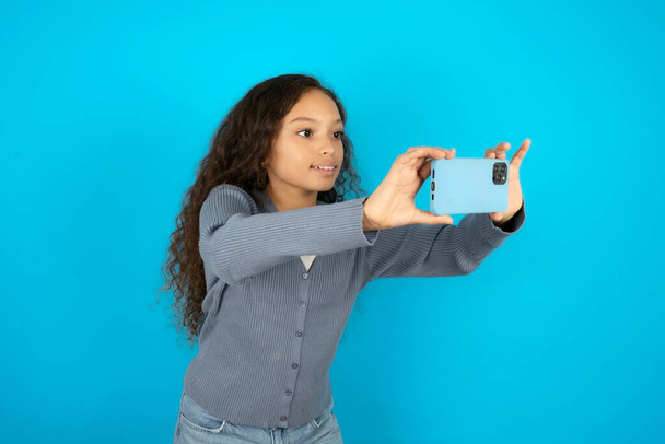 Beautiful kid girl wearing casual jacket over blue background taking a selfie to post it on social media or having a video call with friends. - Photo, Image