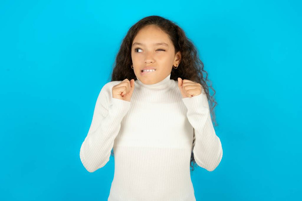 Beautiful kid girl wearing white turtleneck over blue background clenches fists and awaits for something nice happened looks away bites lips and waits announcement of results - Foto, afbeelding