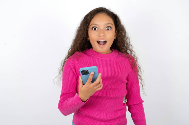 Красивая девочка в розовой водолазке на белом фоне держит в руках мобильный телефон и радуется позитивным новостям, использует современные сотовые - Фото, изображение