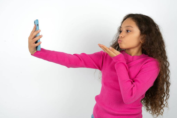 Hermosa niña con cuello alto rosa sobre fondo blanco sopla beso de aire en la cámara del teléfono inteligente y toma selfie, envía mwah a través de llamada en línea. - Foto, imagen