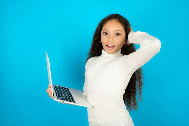 Photo of amazed beautiful kid girl wearing white turtleneck over blue background holding modern gadget arm head - Photo, Image