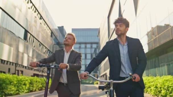 Бизнес-менеджеры держат скутер-велосипед в центре города. Корпоративные люди ездят пешком в современном офисном здании. Успешные коллеги в костюмах с использованием эко-транспорта утром - Кадры, видео