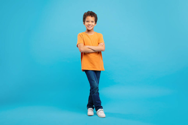 Впевнений молодий школяр з доброзичливою посмішкою, схрещеними руками, одягнений в помаранчеву футболку та джинси на заспокійливому синьому фоні, повна довжина - Фото, зображення