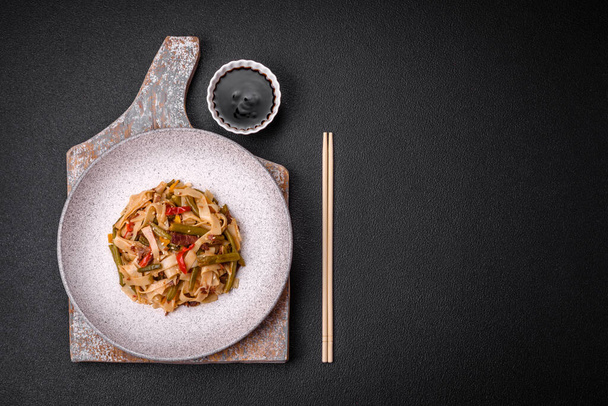 Köstliches asiatisches Gericht Udon mit Paprika, grünen Bohnen, Gewürzen und Kräutern auf einem Keramikteller auf dunklem Betongrund - Foto, Bild