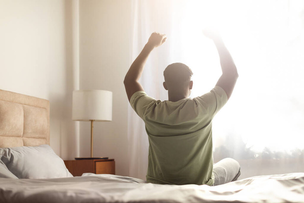 Вид сзади на отдохнувшего неузнаваемого черного парня, сидящего на кровати, растягивающего тело и руки после пробуждения утром, смотрящего на окно спальни с солнечными вспышками. Концепция здорового сна - Фото, изображение