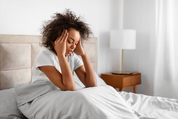 Matin Mal de tête. Malheureuse jeune femme noire assise au lit et touchant sa tête ayant des douleurs migraines, lutte contre les maux de tête chroniques dans l'intérieur de la chambre moderne - Photo, image