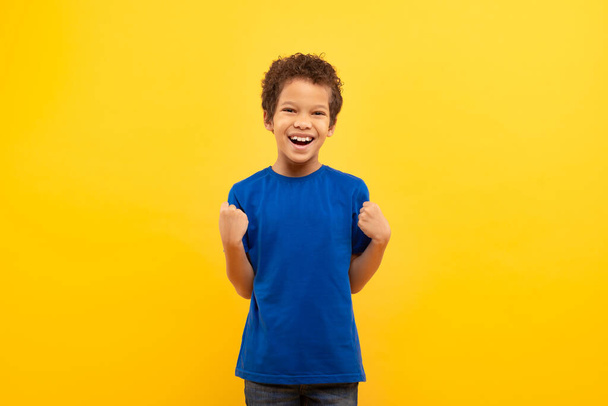 Nadšený chlapec s kudrnatými vlasy, smějící se a zaťaté pěsti ve vítězném póze, slaví úspěch, nosí královsky modré tričko na žlutém pozadí - Fotografie, Obrázek
