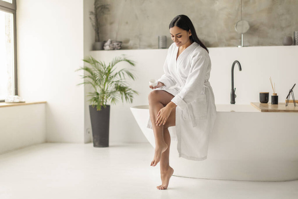 Junge langhaarige Indianerin im weißen Bademantel, die Körperpeeling oder Creme auf die Haut der Beine aufträgt, im Luxus-Badezimmer sitzend, leere Kopierfläche für Werbung. Körperpflegekonzept - Foto, Bild