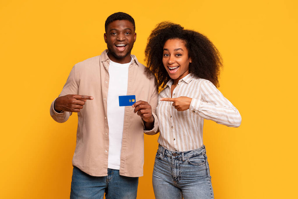 Uśmiechnięta młoda, czarna para z podnieceniem wskazująca na posiadaną kartę kredytową, sugerująca finansową aprobatę lub nowy zakup, na słonecznym żółtym tle - Zdjęcie, obraz