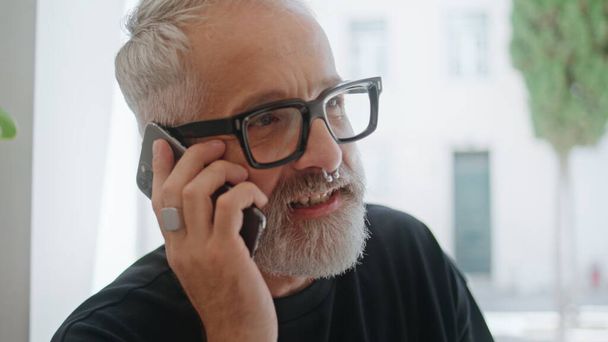 Stylish man chatting smartphone closeup. Handsome senior ending call working alone. Smiling bearded designer talk mobile phone with client. Good looking mature male wearing piercing enjoy conversation - Photo, Image