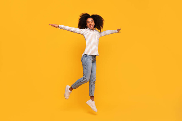Zářící mladá černoška s rukama široce otevřenýma radostně skáče proti hořčičně žlutému pozadí, sportovní ležérní elegantní s pruhovanou košilí a džínami - Fotografie, Obrázek