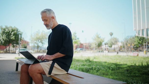 Stílusos végzős gépelő számítógép a városi parkban. Figyelmes ember, aki kint pihen a szabadban, és fülhallgatóban böngészi az internetet. Jóképű, szakállas férfi, szörf laptopot készít. A nyugdíjba vonulás fogalma. - Fotó, kép