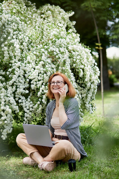 Boldog fiatal nő beszél telefonon, laptopot használ fehér virágzó virágokban. Távmunka, csevegés barátokkal, koncepció tanulás. - Fotó, kép