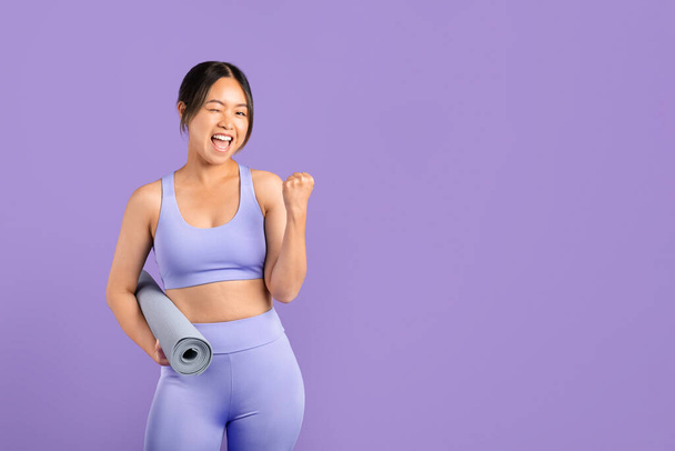 Joven mujer asiática en ropa deportiva tiene alfombra de fitness y gestos entusiastas sí, transmitiendo positividad y determinación sobre el fondo púrpura, espacio libre - Foto, imagen