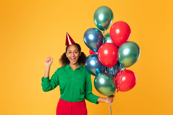 Happy African American teini tyttö juhlava syntymäpäivä hattu poseeraa joukko värikkäitä ilmapalloja, eleiden kyllä juhlii hänen erityinen päivä yli keltainen studio tausta - Valokuva, kuva