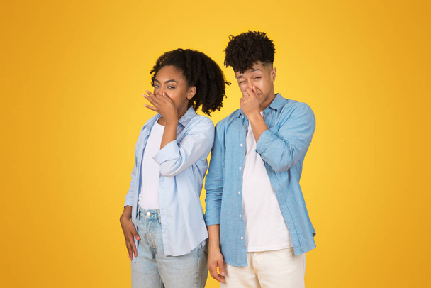 Két fiatal fekete alkalmi öltözékben játékos gesztussal takarta el a száját, egy titokra vagy meglepetésre utalva, élénk sárga háttér előtt állva, szórakoztató kifejezésekkel. - Fotó, kép