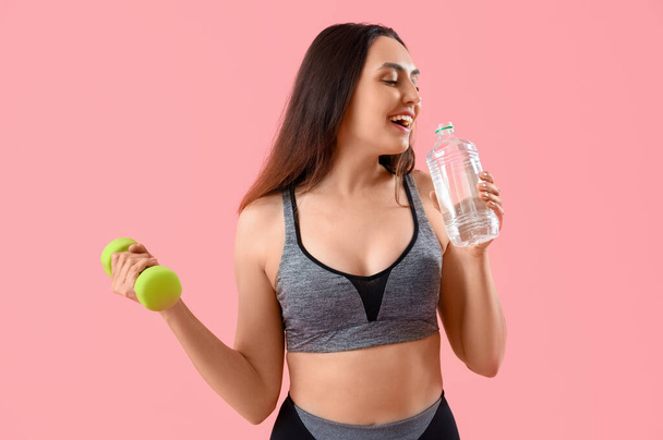 Giovane donna sportiva con bottiglia d'acqua e manubri su sfondo rosa - Foto, immagini