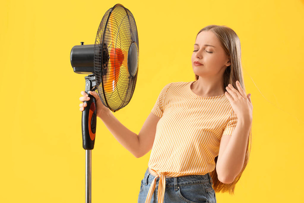 Verschwitzte junge Frau mit elektrischem Ventilator auf gelbem Hintergrund - Foto, Bild