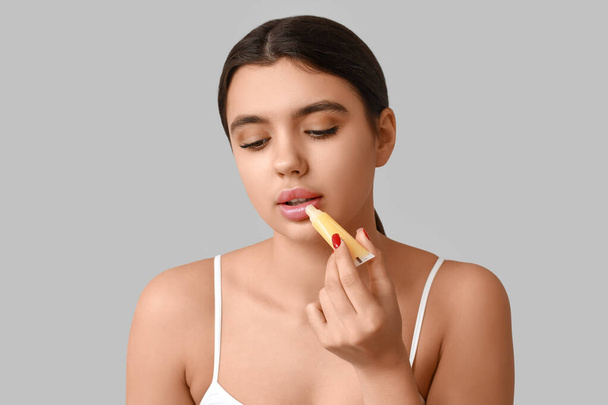 Όμορφη νεαρή γυναίκα εφαρμογή lip balm σε γκρι φόντο - Φωτογραφία, εικόνα