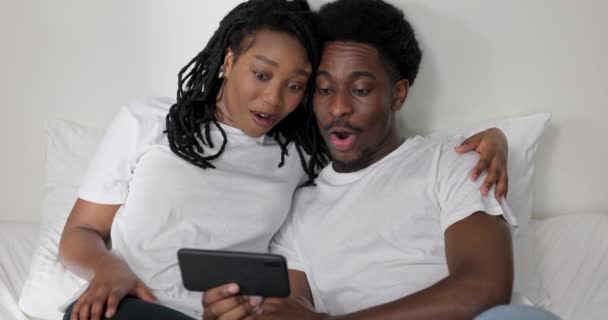 Щаслива американська темношкіра жінка і чоловік, сидячи на ліжку посміхаючись, тримає мобільний телефон в руках, дивлячись новини фільми телепередачі посміхаючись сміється обіймаючись в сучасній затишній спальні. - Кадри, відео