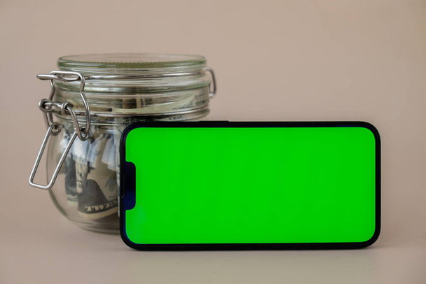 horizontal Green screen on modern mobile phone in background of glass jar full of American currency dollar banknotes on beige background. Cope space for text. Advertisement for application website - Photo, Image
