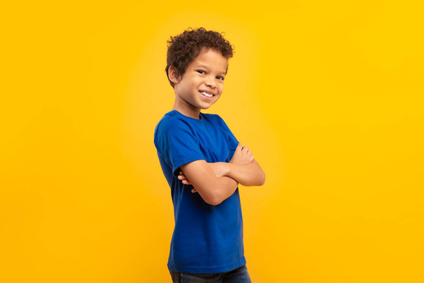 Sebevědomý latinskoamerický chlapec s kudrnatými vlasy a širokým úsměvem, ruce zkřížené v modrém tričku, stojící na živém žlutém pozadí - Fotografie, Obrázek