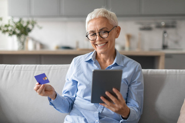 E-Commerce. Ältere graue Dame kauft online ein, bezahlt digital per Tablet-Computer, hält Kreditkarte, während sie etwas im Webshop kauft, sitzt auf dem Sofa im modernen Wohnbereich - Foto, Bild