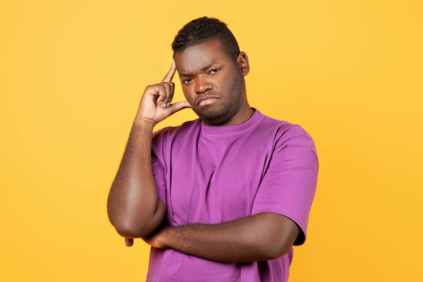 Στοχαστικός δυσαρεστημένος μαύρος που σκέφτεται τα προβλήματά του Αγγίζοντας το κεφάλι, κοιτάζοντας την κάμερα με ψυχική έκφραση, στέκεται πάνω από το κίτρινο φόντο στούντιο, φορώντας μωβ T-shirt - Φωτογραφία, εικόνα