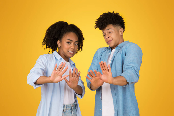 A cheerful young black woman and man stand back to back against a vibrant yellow background, smiling and playfully gesturing stop with their hands up in a casual and fun pose - Photo, Image